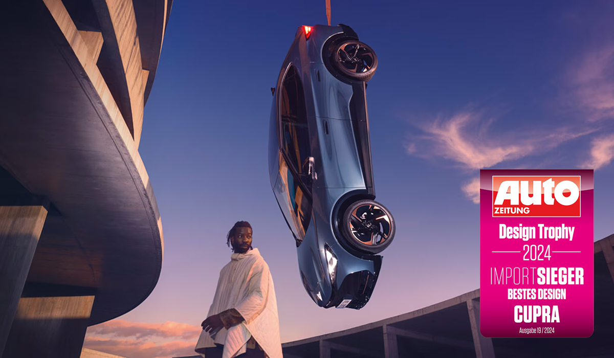 CUPRA Tavascan in Frontansicht auf nasser Fahrbahn vor Berglandschaft mit Design Trophy Plakat im Vordergrund